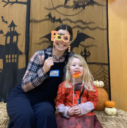 A woman and a girl in costumes pose with playful props for Halloween, surrounded by pumpkins and a spooky backdrop.