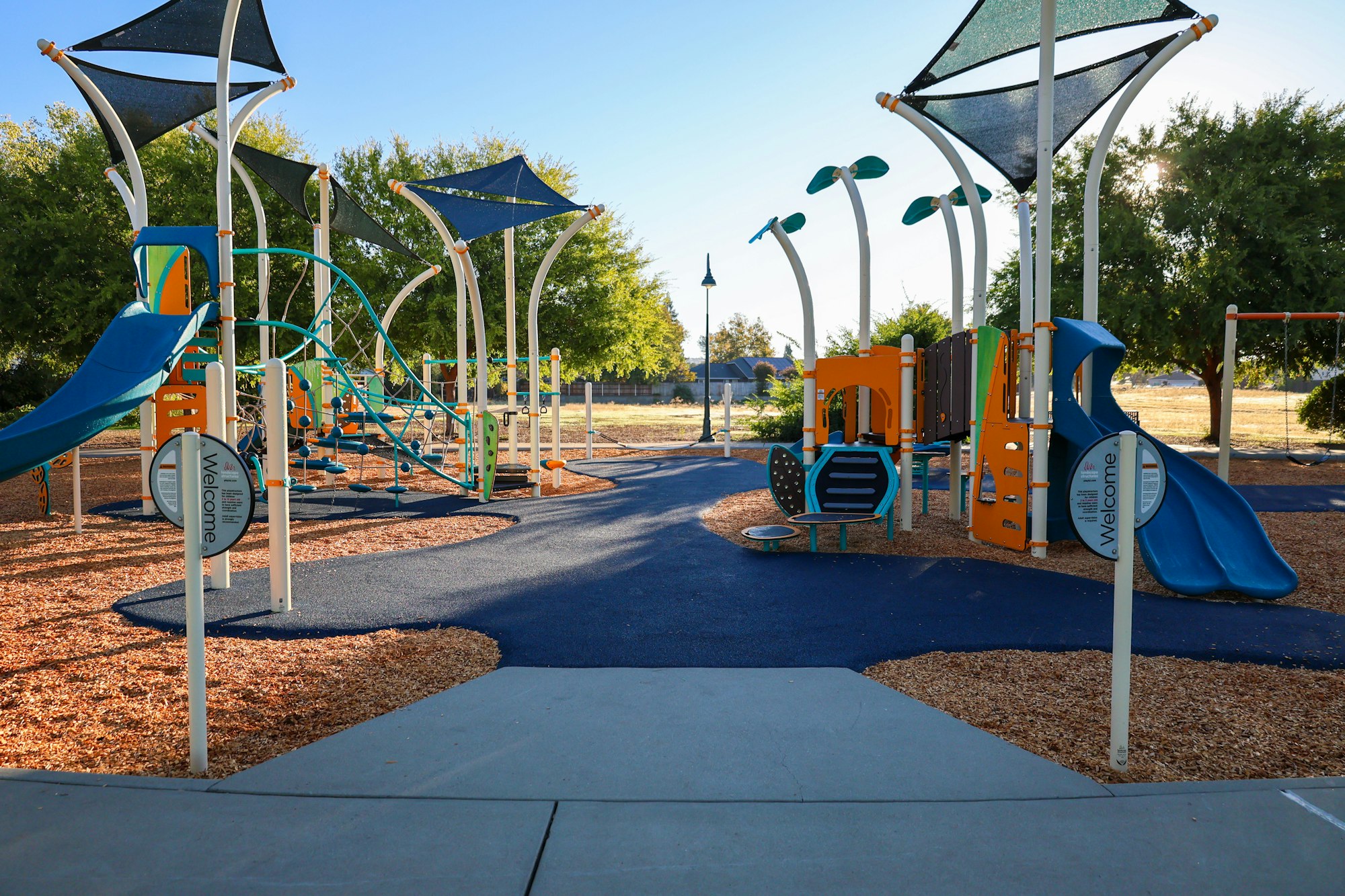 A colorful playground with slides, climbing structures, shade canopies, and wood chip ground covering.