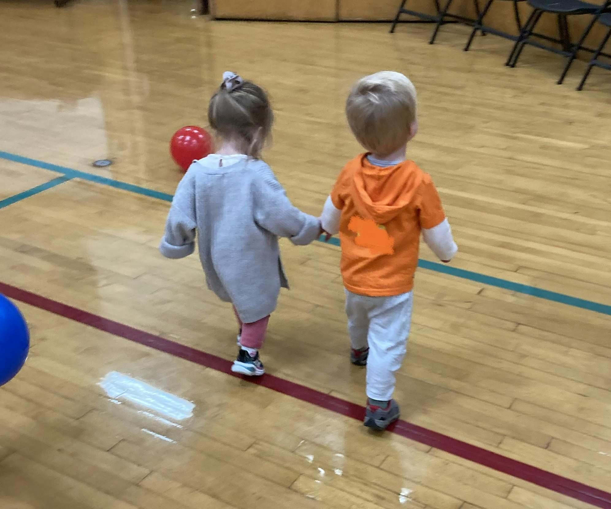 Two young children holding hands in a gymnasium, with a red and a blue ball nearby.
