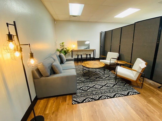 A cozy room with a gray sofa, armchairs, round coffee table, lamps, plants, and a patterned rug on wooden flooring.