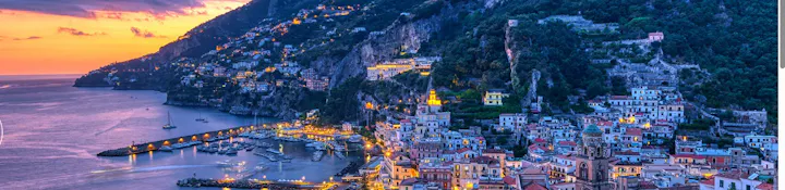 A scenic coastal town at dusk, featuring colorful buildings, a marina, and illuminated cliffs along the shoreline.