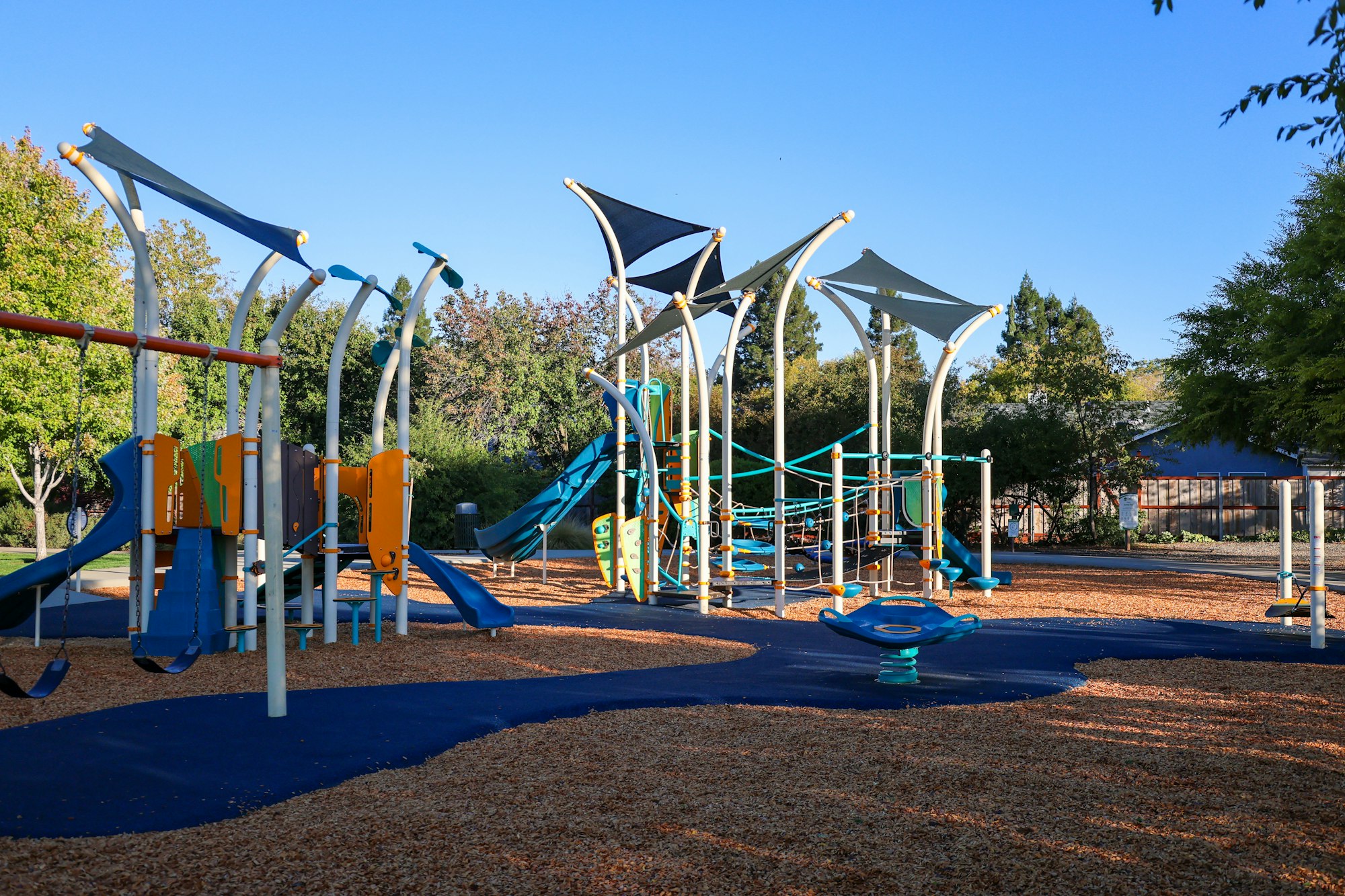 Colorful playground with slides, swings, and climbing structures on a sunny day, surrounded by trees.