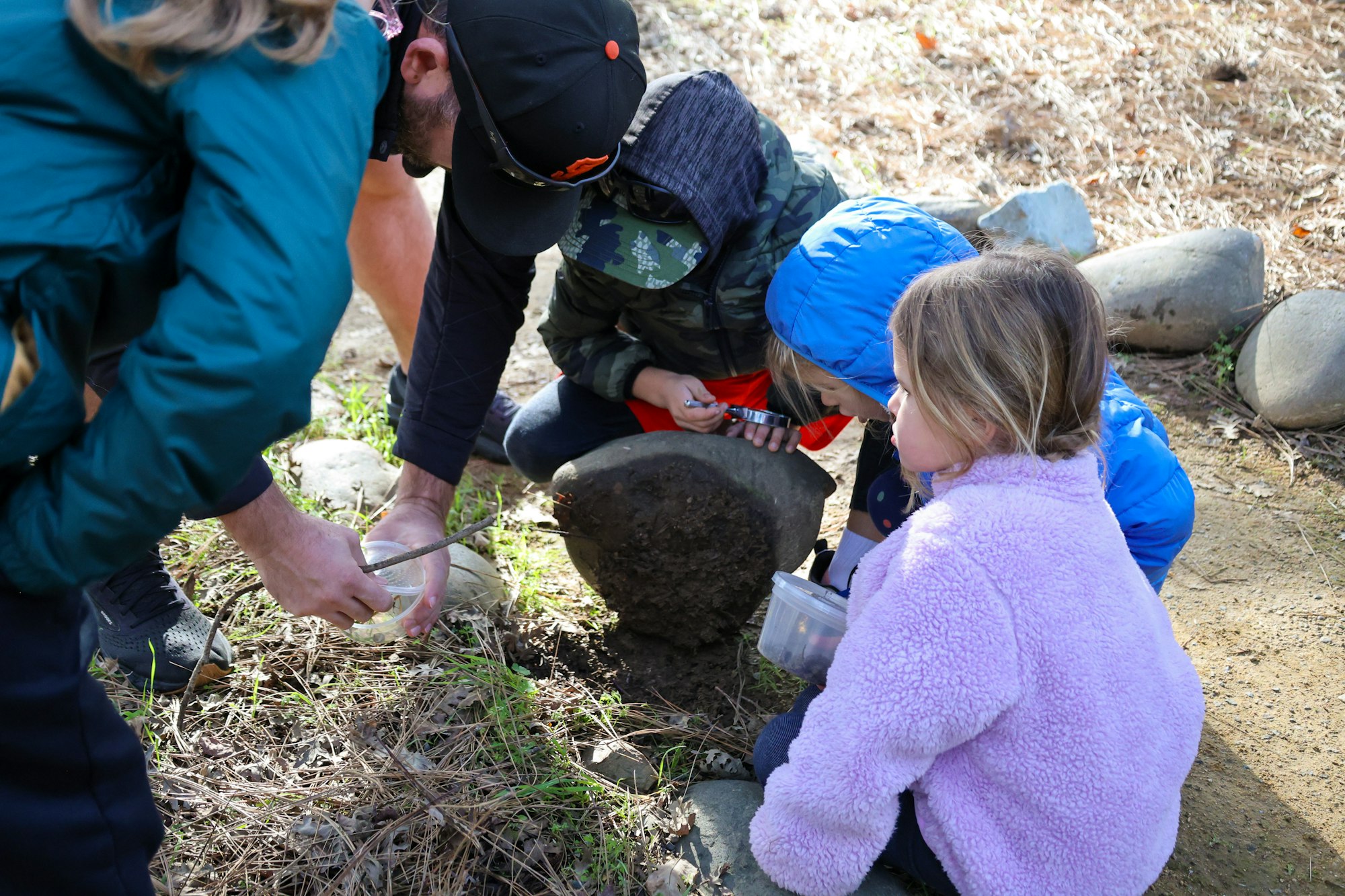 A group of children and an adult explore nature, examining objects and using tools near a rock and grassy area.