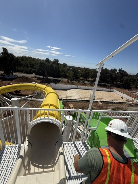 An elevated view of a yellow water slide under construction, with a worker in a hard hat and safety vest nearby.