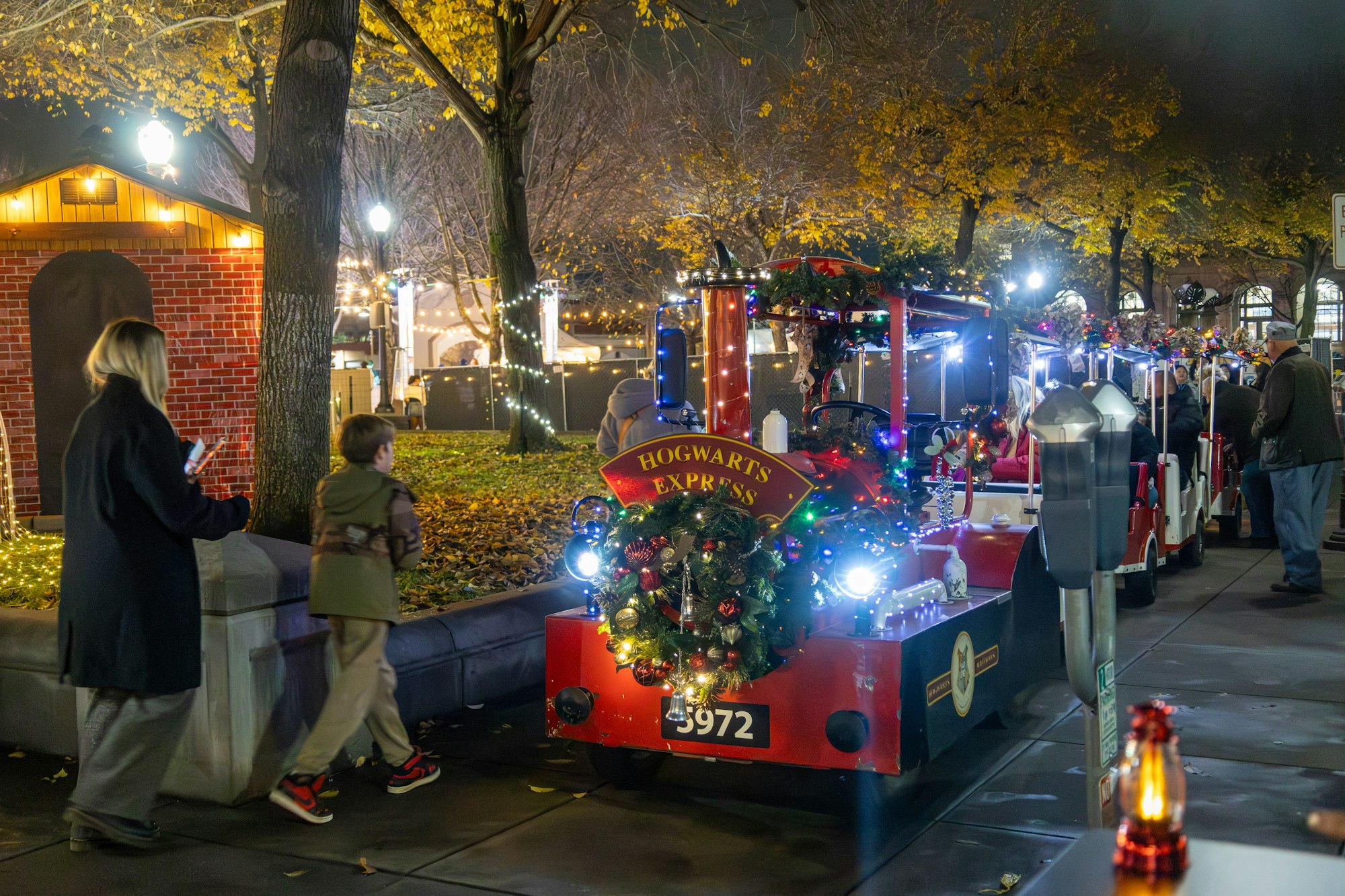 A festive Hogwarts Express train with decorations, surrounded by people and colorful lights in an outdoor setting at night.