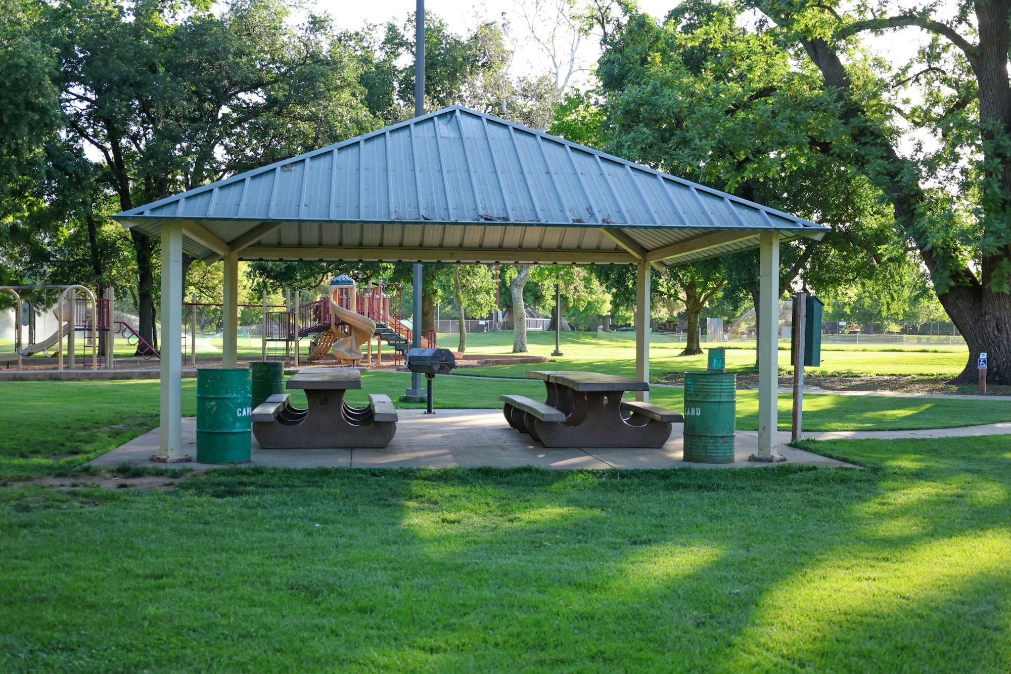 A park with a covered picnic area, benches, trash cans, and a playground in the background, surrounded by trees.