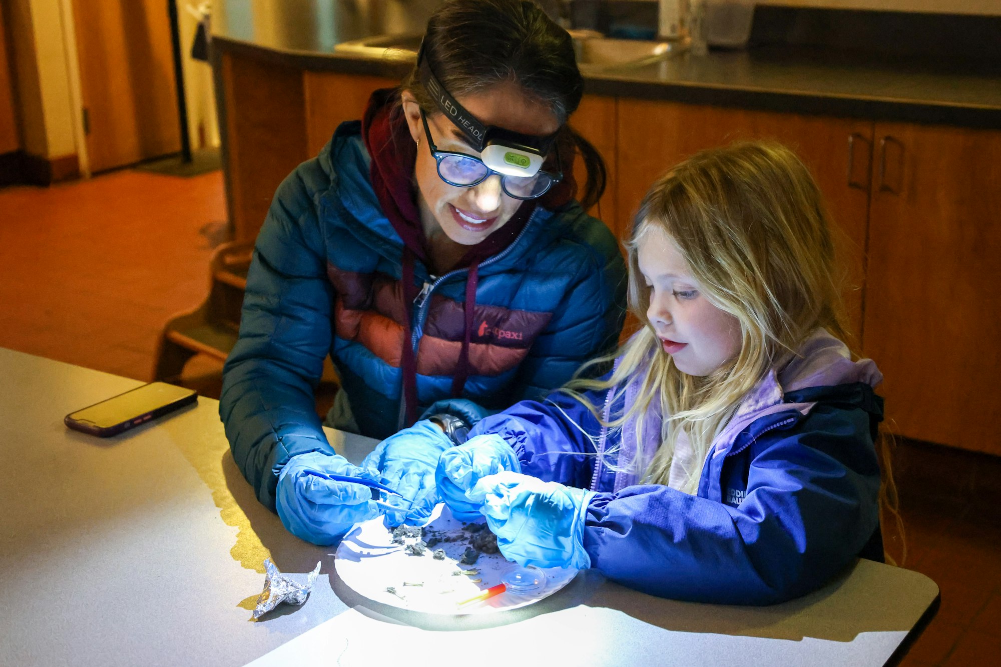 Two people in jackets and gloves examine objects under a light, using tweezers on a table.