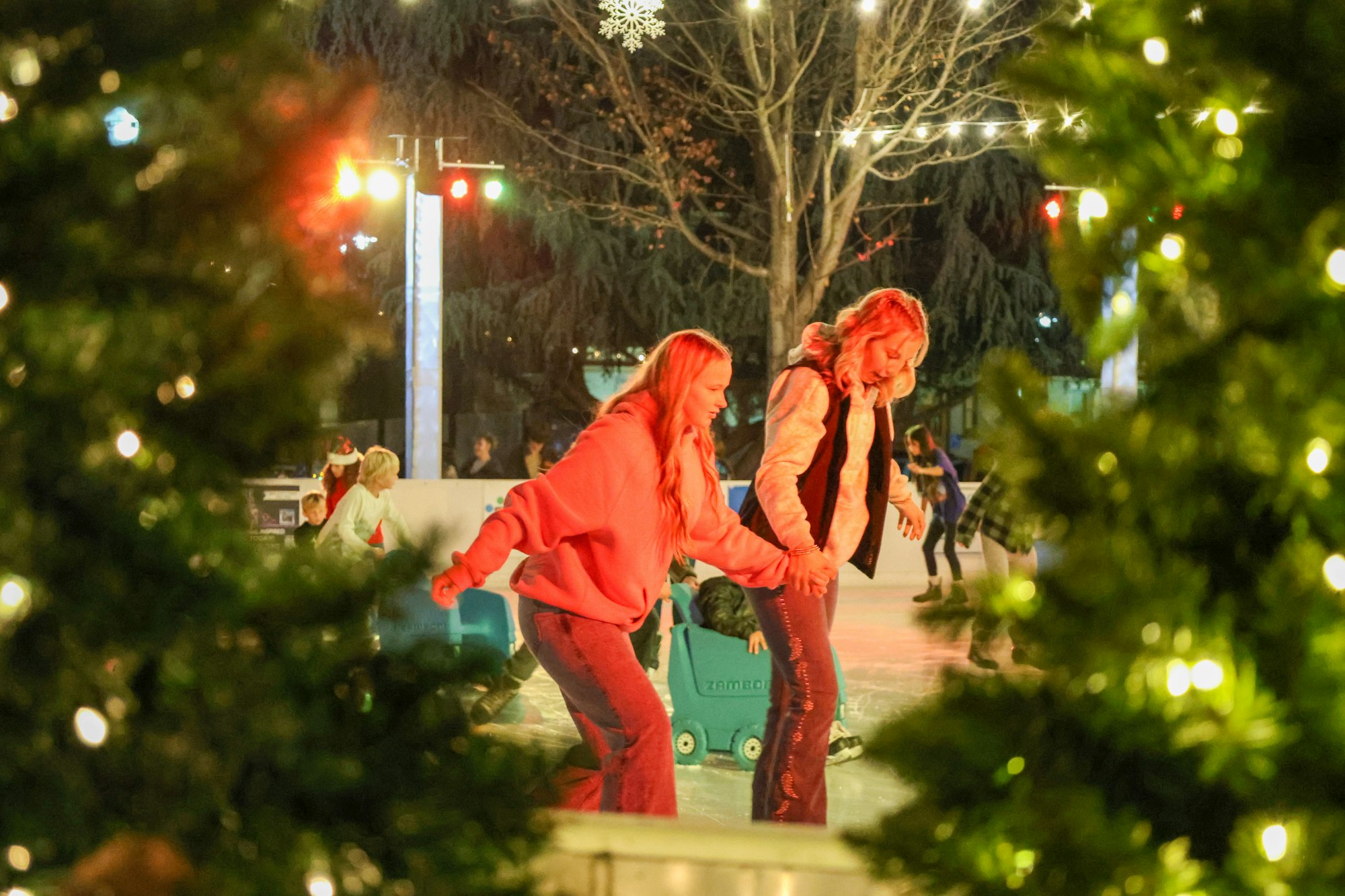 Two young women ice skating together at a festive outdoor rink, surrounded by holiday decorations and twinkling lights.