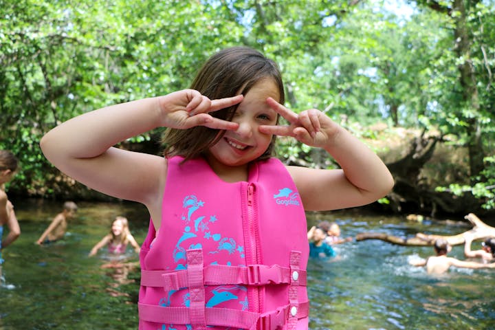 Child in pink life jacket making peace signs at a creek with others swimming in the background.