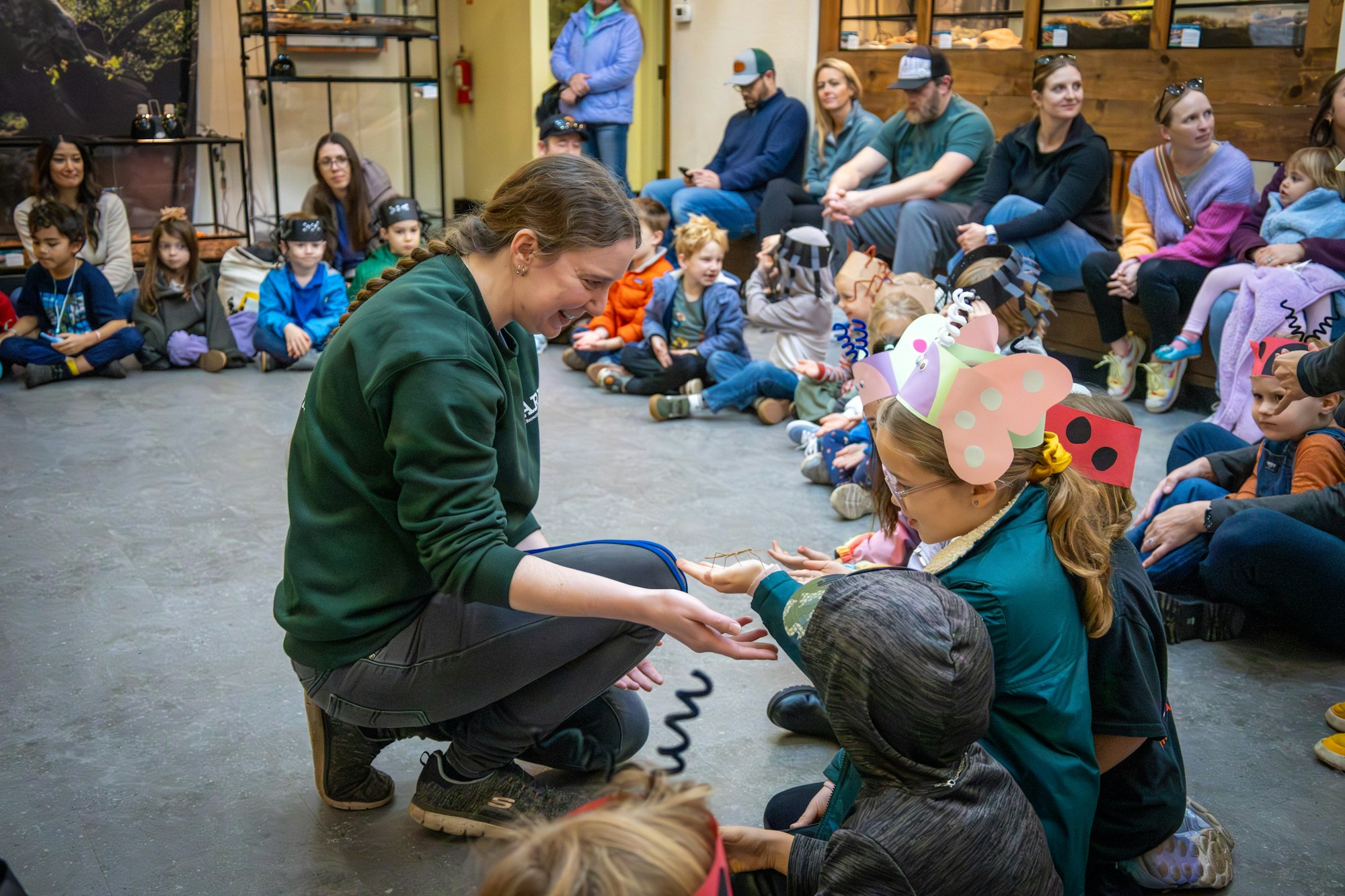 A group of children watches a woman demonstrate holding an insect or small creature during an educational program.