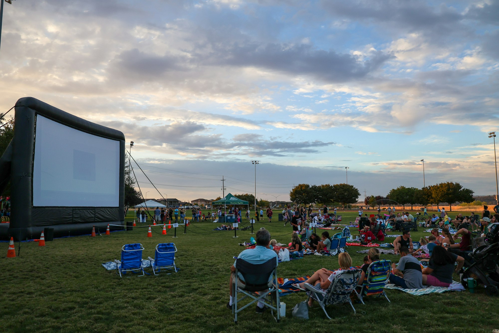 Outdoor movie night with a large inflatable screen and people sitting on the grass with chairs and blankets.