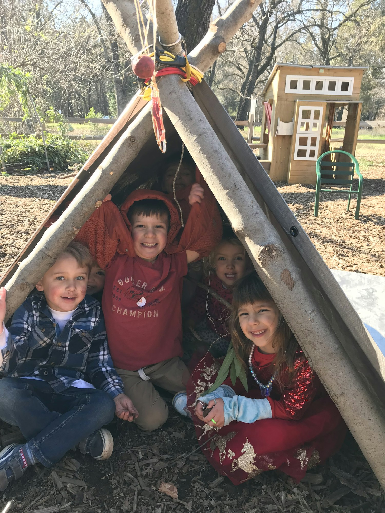 Four children smiling inside a makeshift wooden shelter outdoors with a playhouse visible in the background.