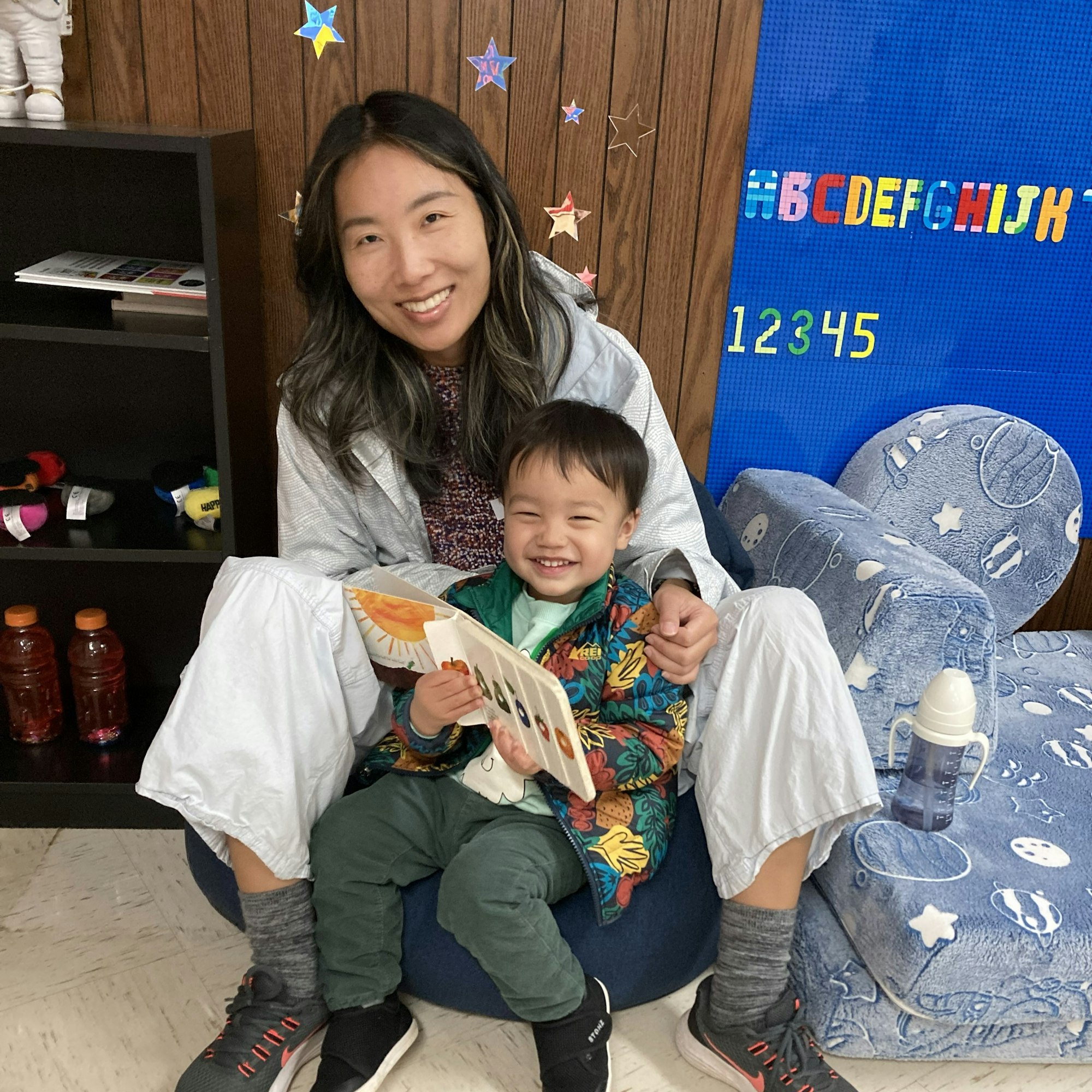 A woman and child sit together in a cozy, colorful space; the child is holding a book and smiling.