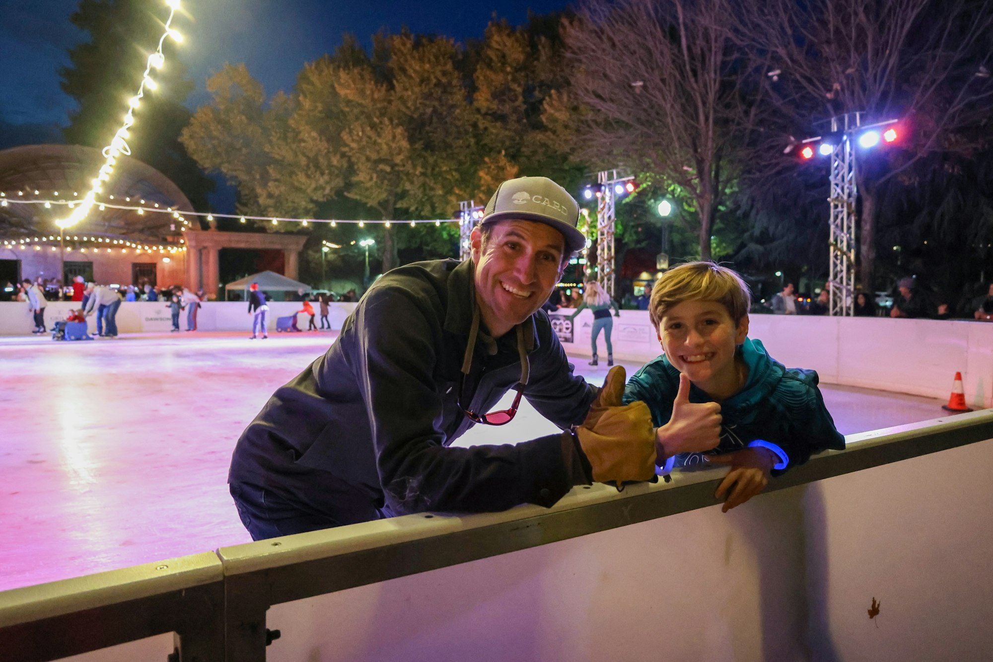 Two people smiling and giving thumbs-up at an ice rink with lights and trees in the background.
