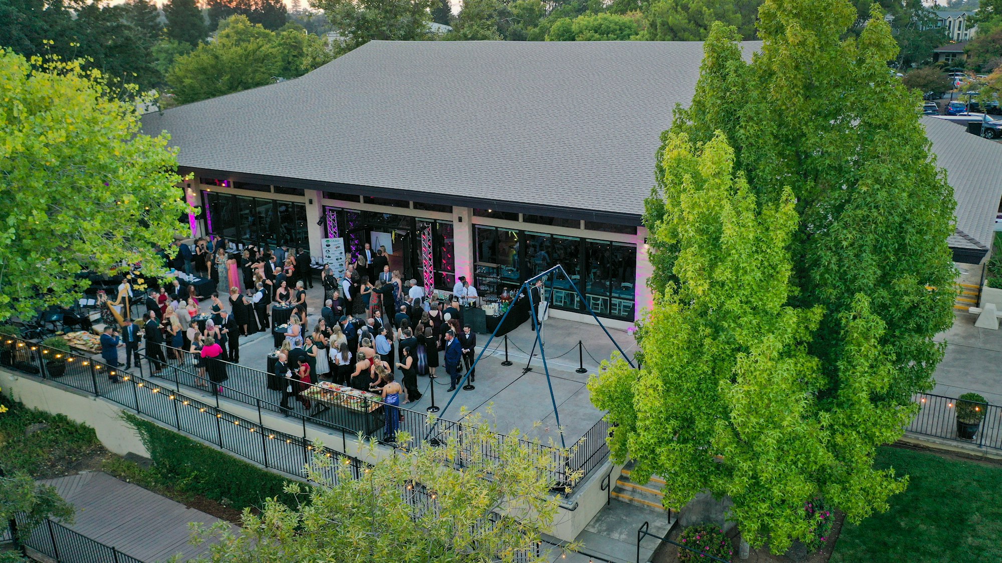 An outdoor event with people in formal attire, set on a large patio surrounded by trees.