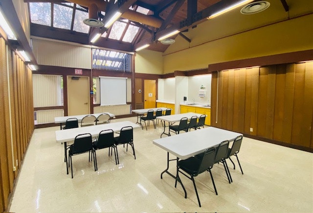 Empty meeting hall with tables, chairs, and a whiteboard. Bright, skylights, and wooden paneling.