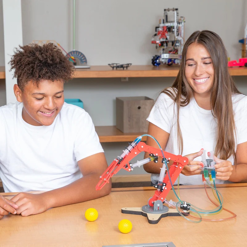 Two kids are smiling while experimenting with a robotic arm that uses syringes to control movement, playing with yellow balls.