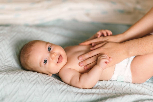 A smiling baby lying down, wearing a diaper, with hands gently touching their chest.
