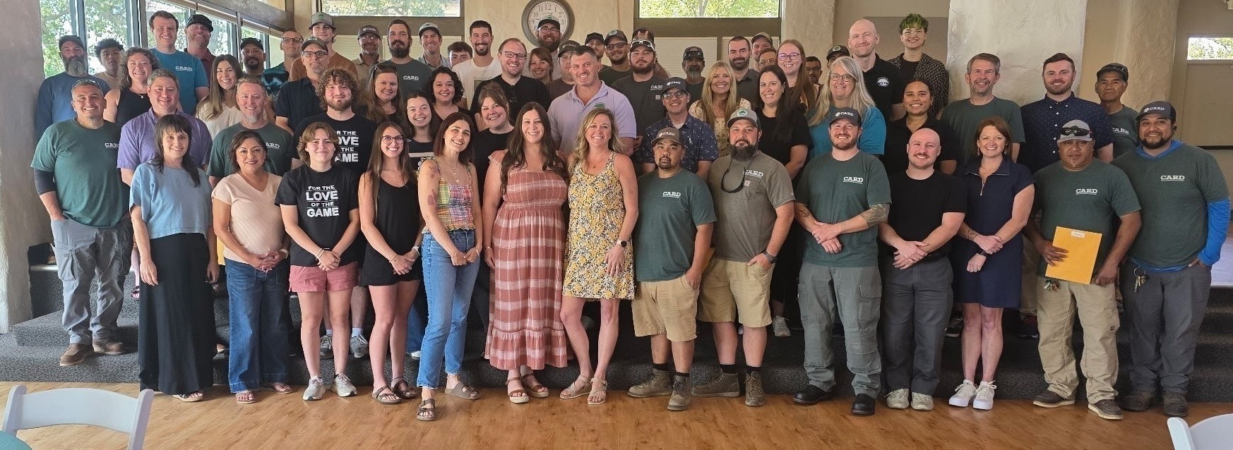 A large group photo of employees, smiling and posing together in a casual setting. Many wear matching shirts.