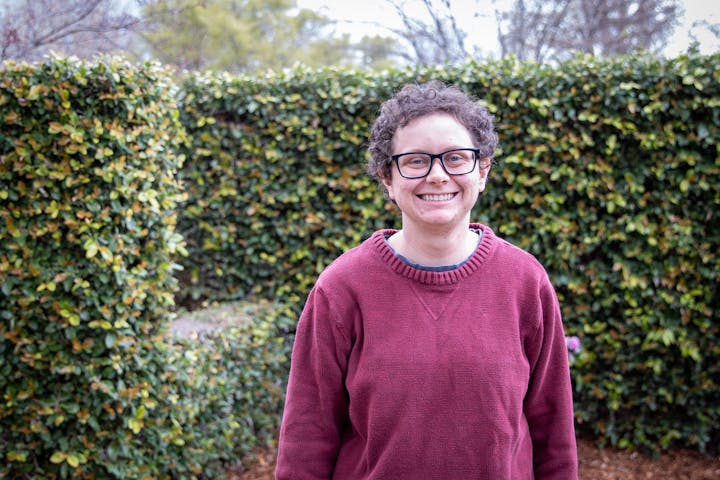 A person wearing glasses and a red sweater smiles in front of a leafy hedge.