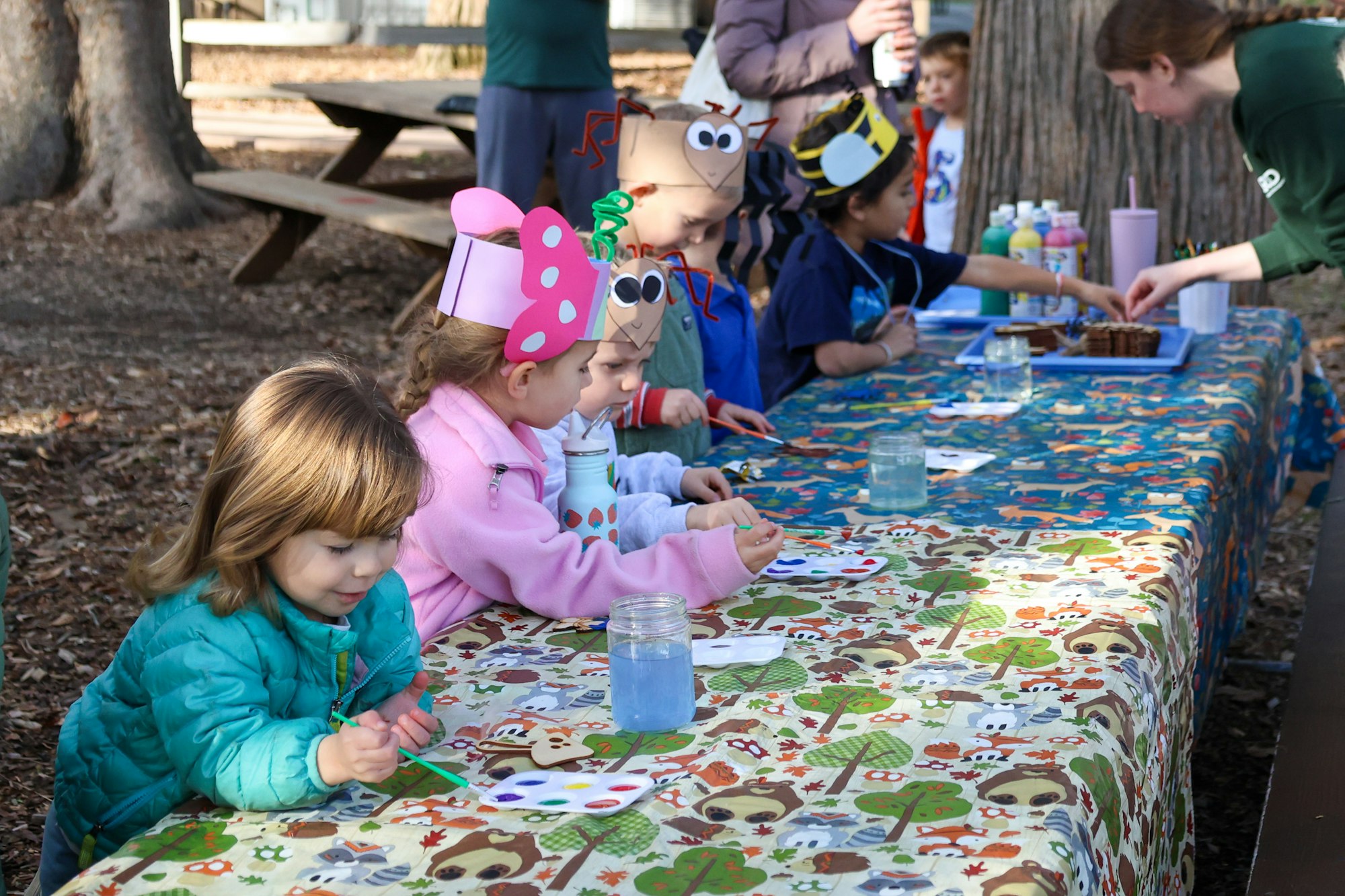 Children are crafting and painting at a picnic table outdoors, wearing fun hats, with artwork supplies and snacks nearby.