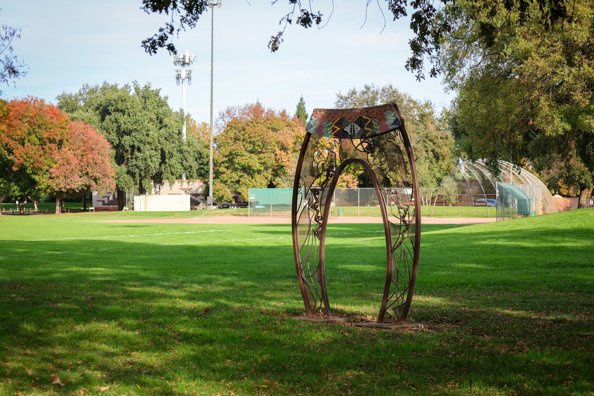 A decorative metal arch stands in a park with green grass, trees, a baseball field, and light poles.