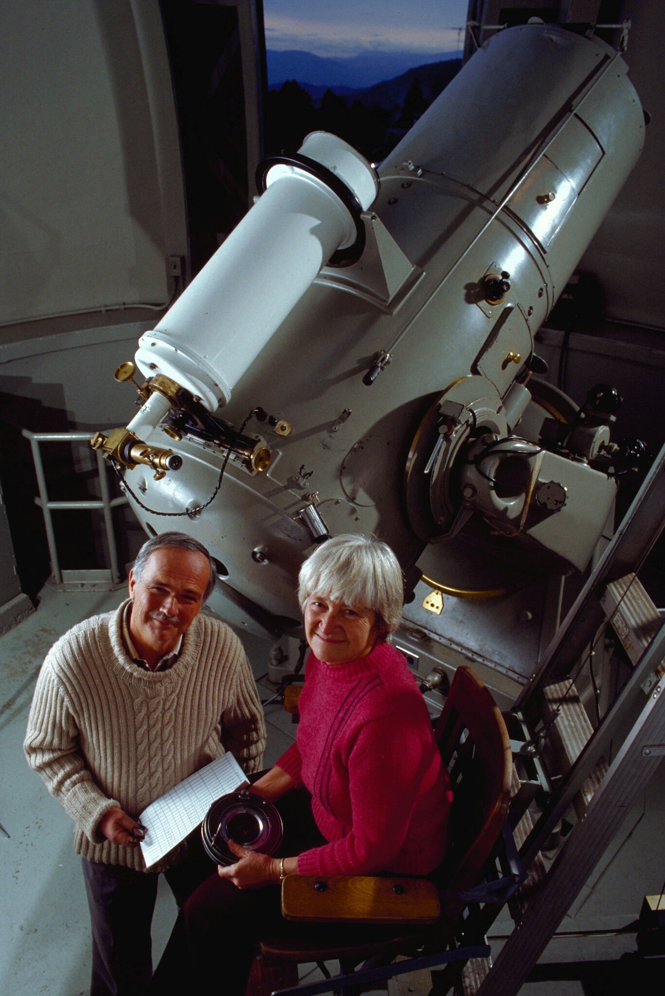 Two people in a telescope observatory, one holding a grid-like sheet, the other holding an object, surrounded by equipment and mountains.