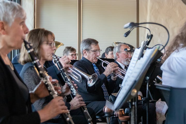 A group of musicians playing brass and woodwind instruments, focused on sheet music on stands.