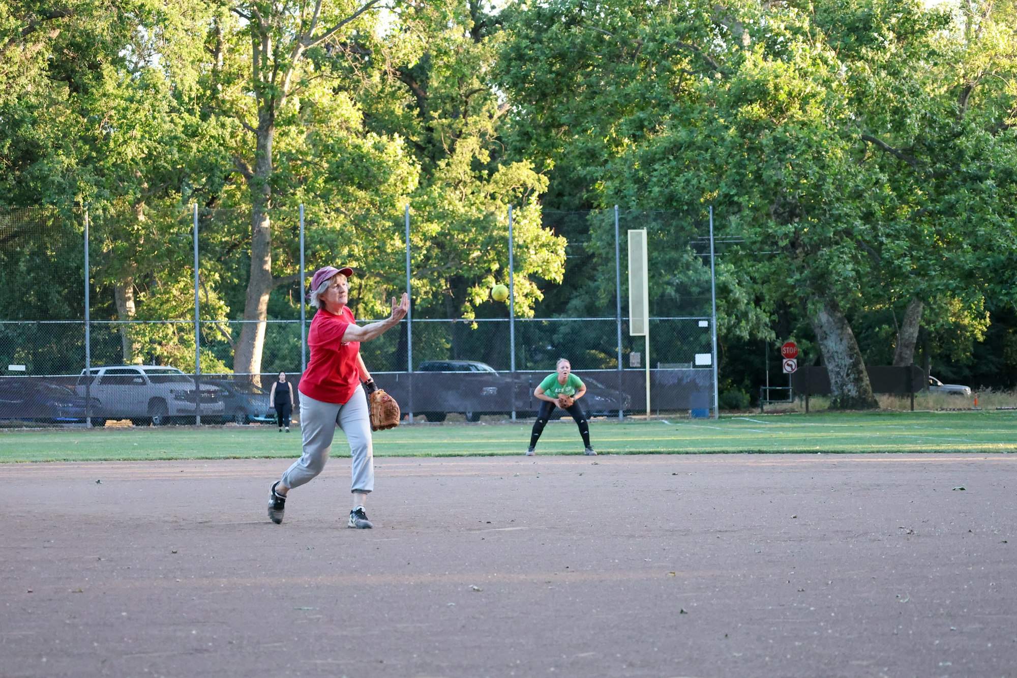 A person in red pitches a softball on a dirt field, watched by another player in green. Trees and parked cars in the background.