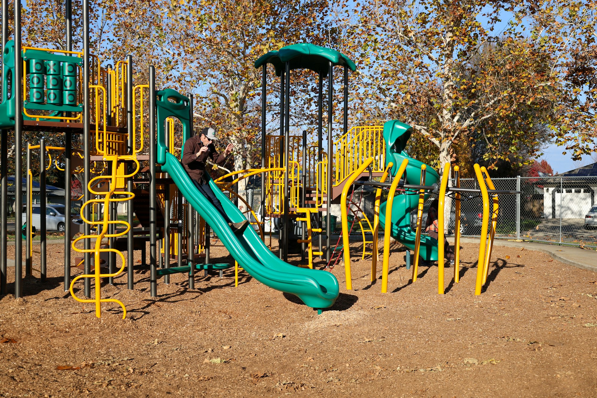A colorful playground with slides and climbing structures, surrounded by autumn trees and a nearby parking area.