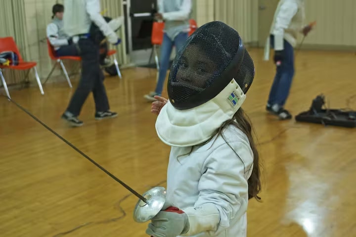 A young fencer in protective gear holds a sword, practicing in a gym with other participants engaged in fencing activities.