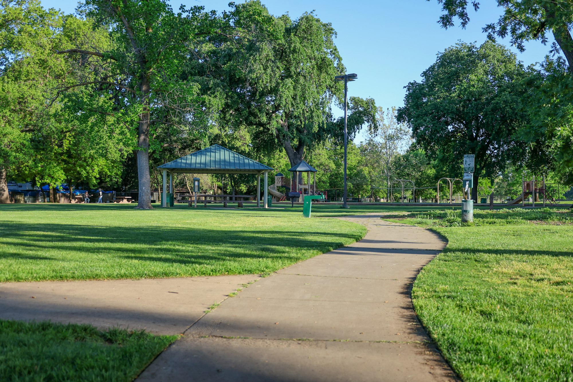 A serene park with green grass, a winding path, picnic tables under a shelter, trees, playground, and a clear blue sky.