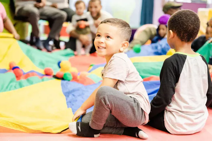 A joyful child sits on a colorful play mat, surrounded by kids and a vibrant parachute during a playful gathering.