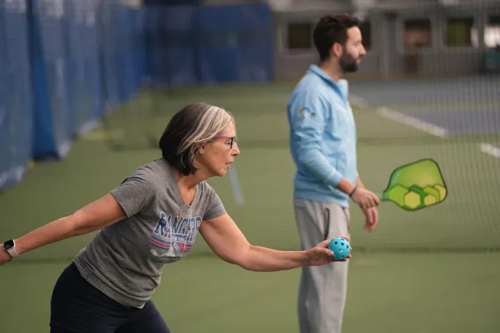 A woman prepares to play with a light blue ball while a man stands by, holding a paddle, in a sports setting.