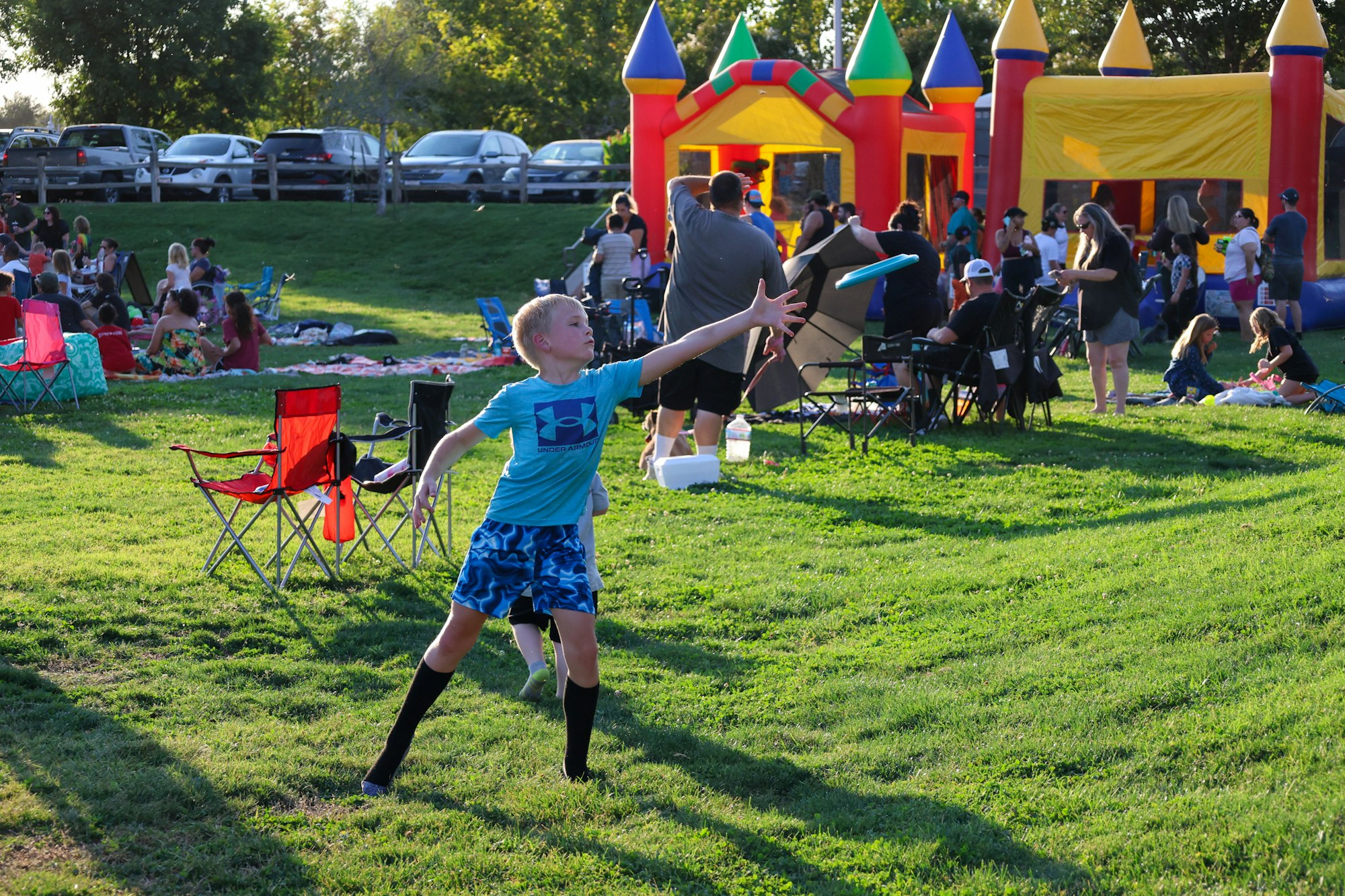 A kid throws a frisbee in a park with people and a bouncy castle around.