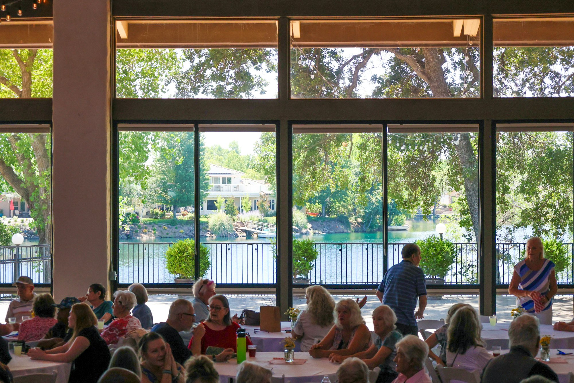 People sitting at tables inside a room with large windows overlooking trees and a lake outside.