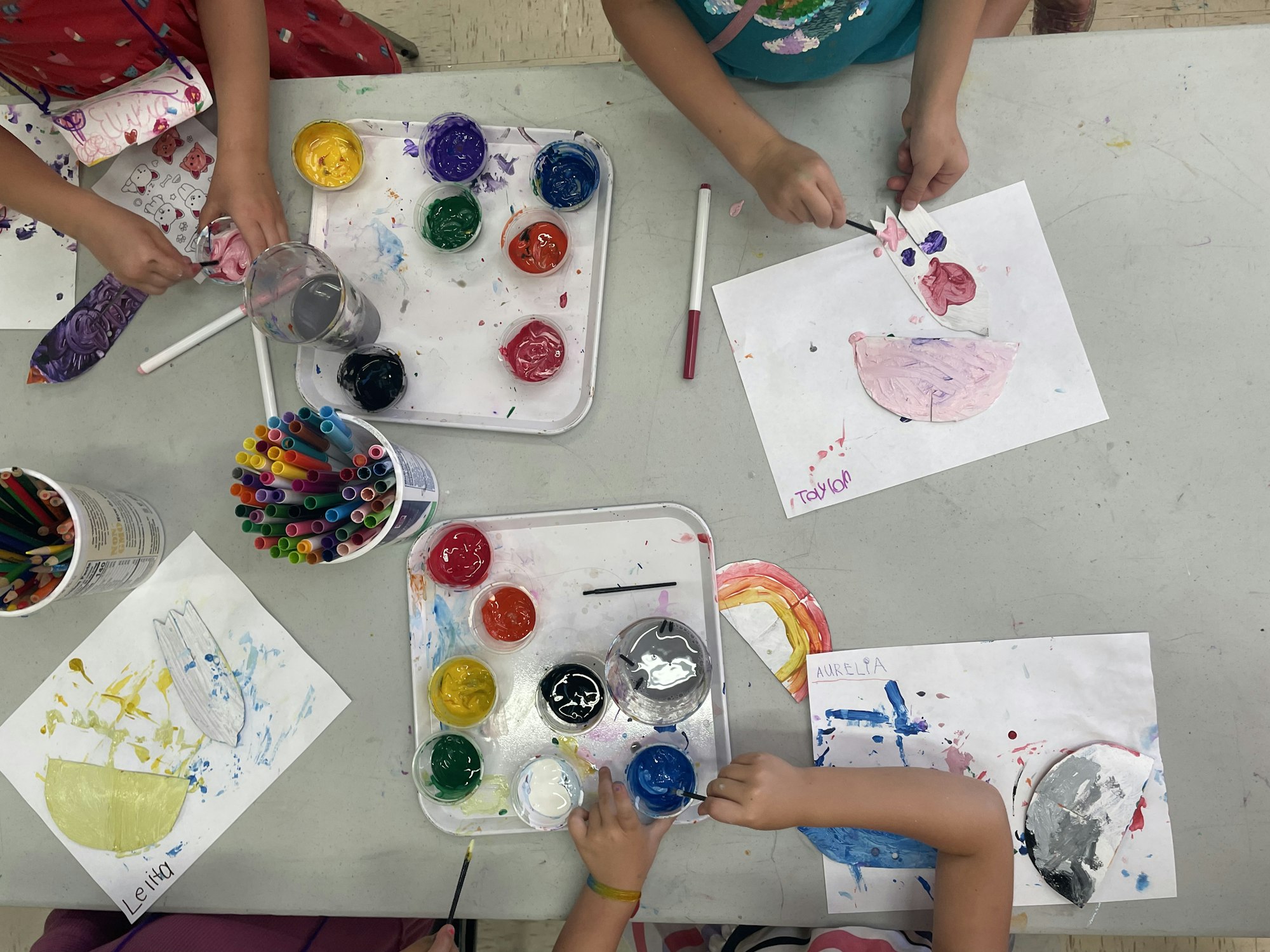 Children are painting and creating colorful art projects with paints, markers, and paper at a creative workspace.