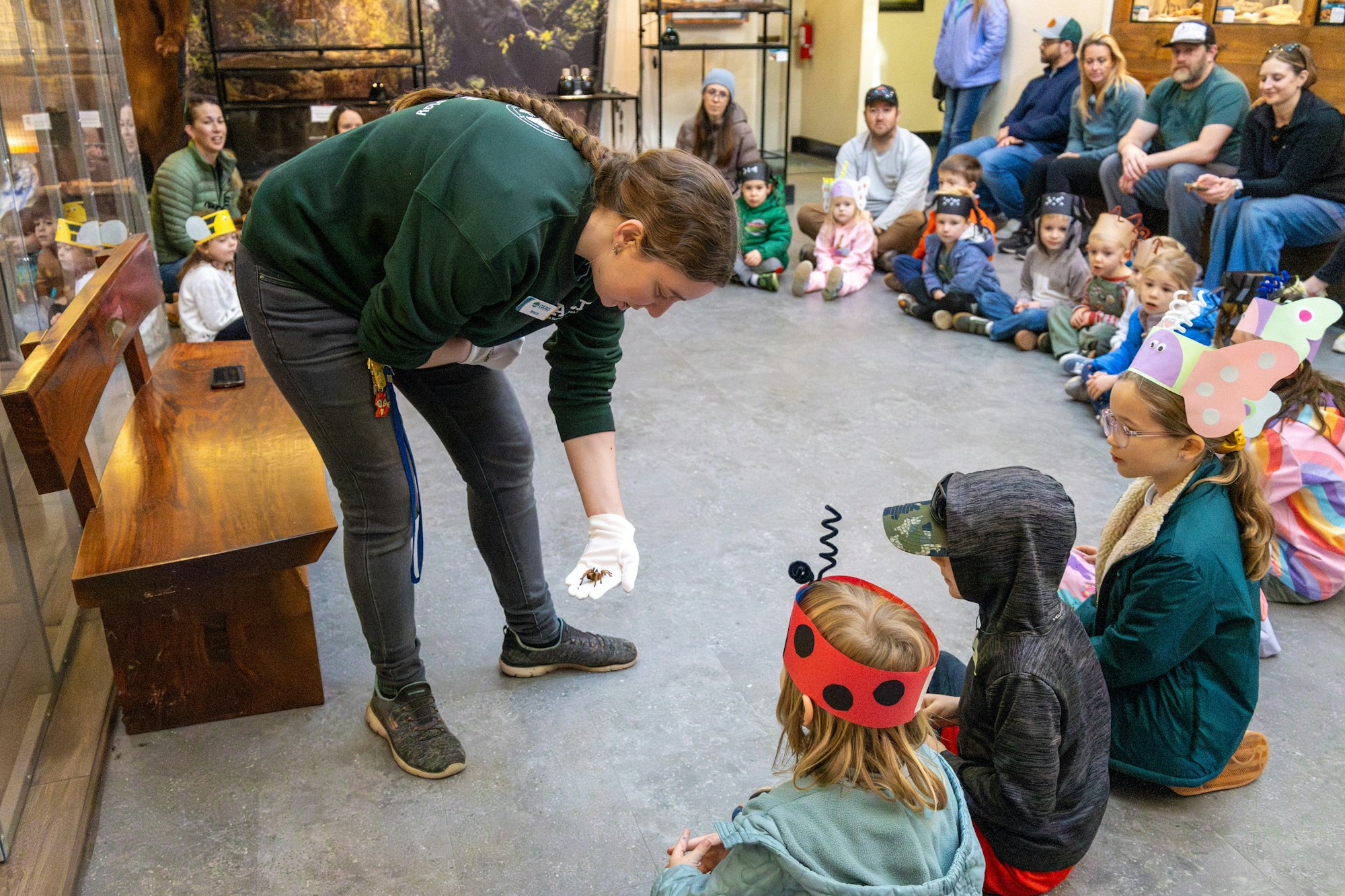 A presenter demonstrates an animal to a group of children, many wearing colorful hats, in an educational setting.