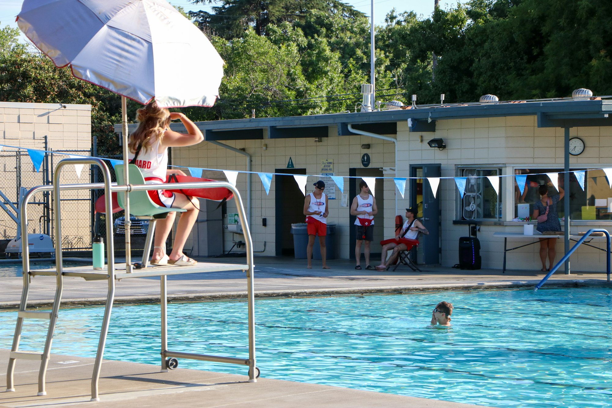 A lifeguard watching over a public swimming pool with people swimming and lounging around.