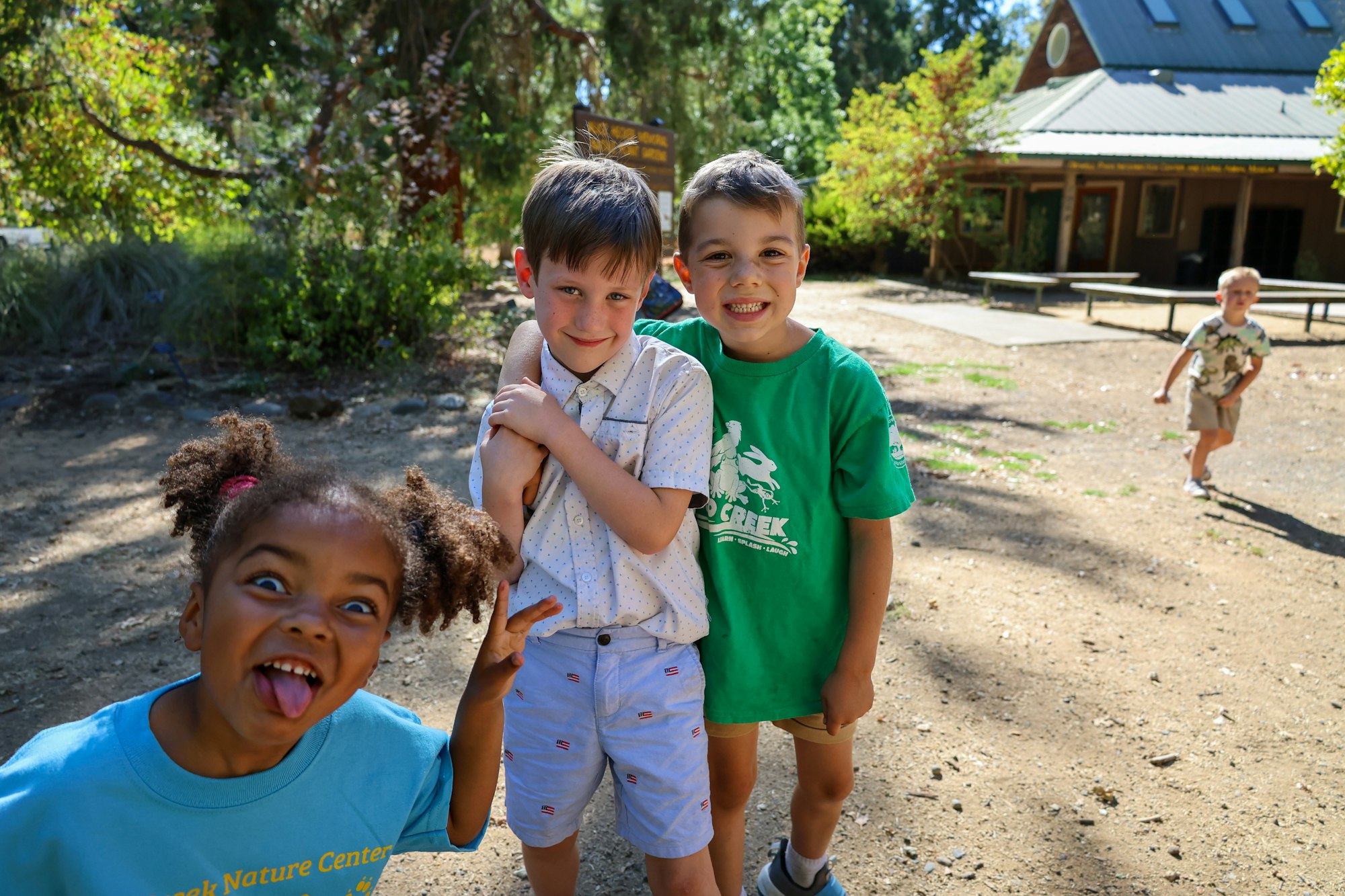 Three children playing and posing outdoors in a sunny park setting. A fourth child is in the background.
