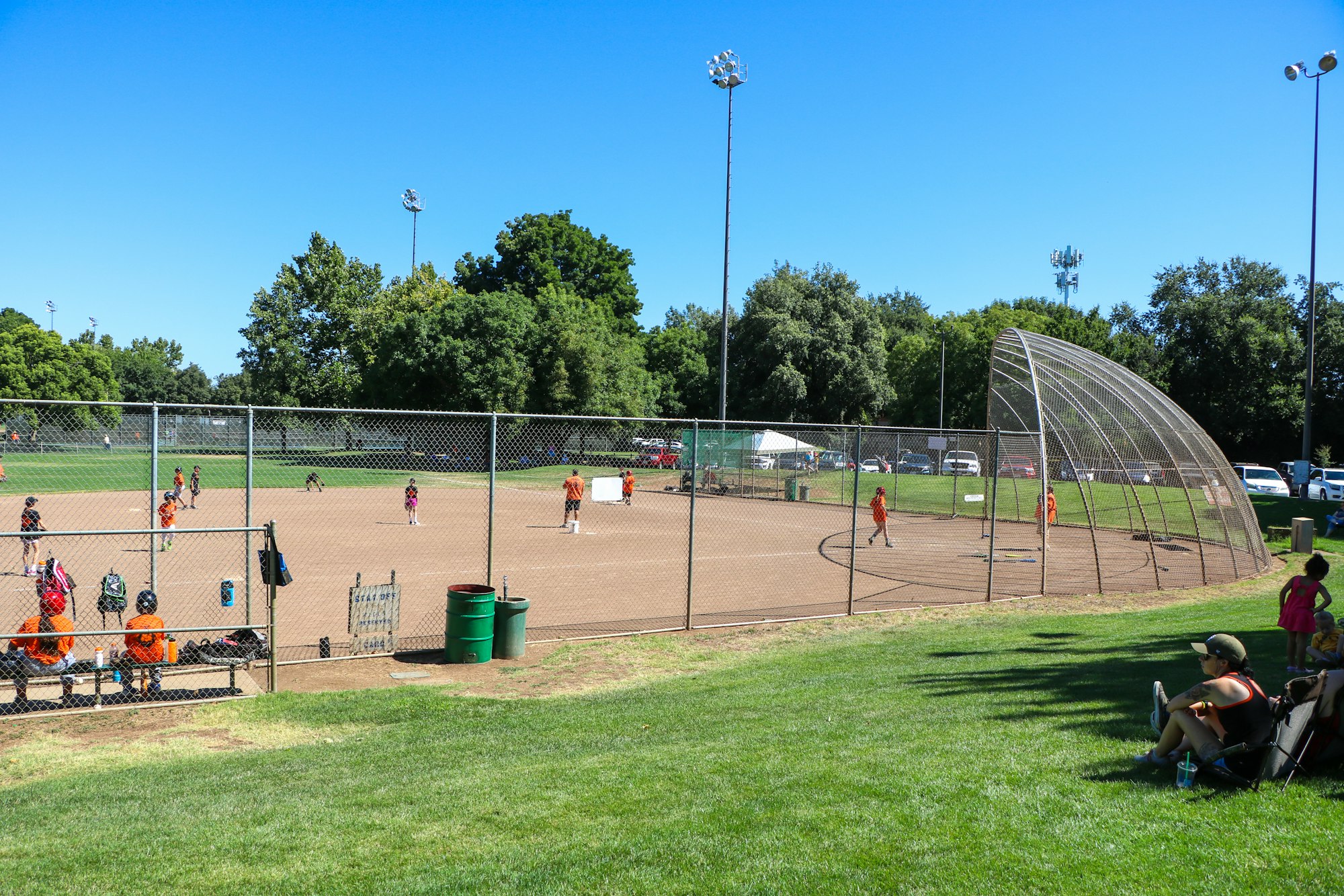 A baseball game in progress on a sunny day, with players on the field and spectators seated on the grass nearby.