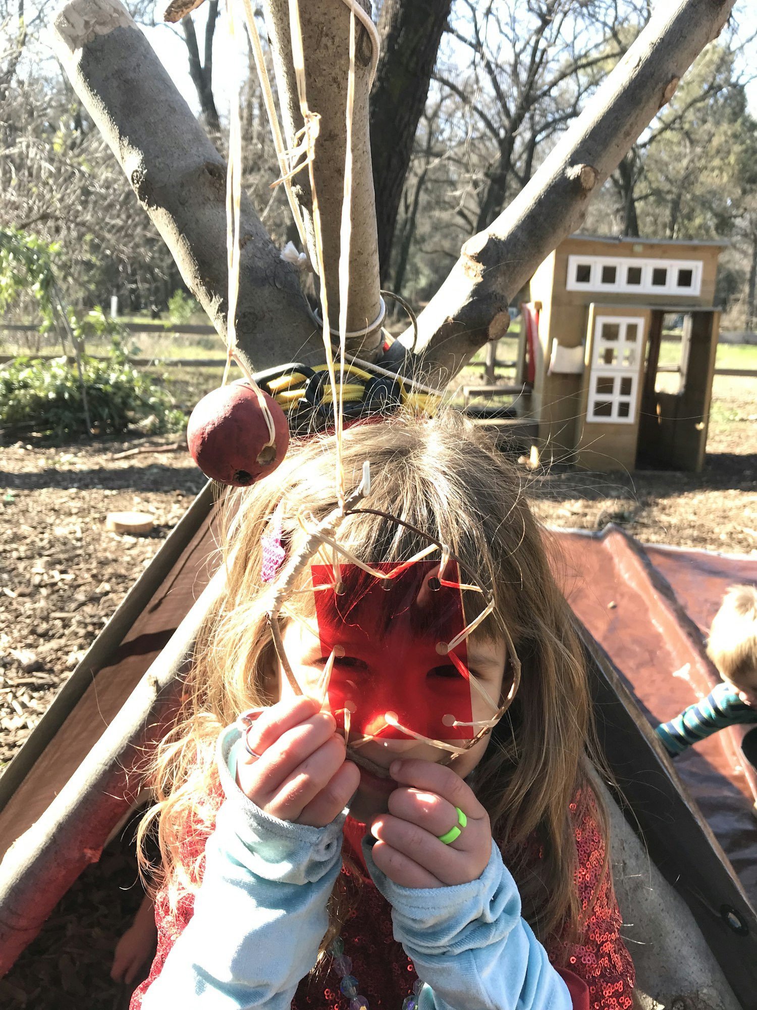 A child holding a red mask outdoors near a playground set with an apple hung from a branch.