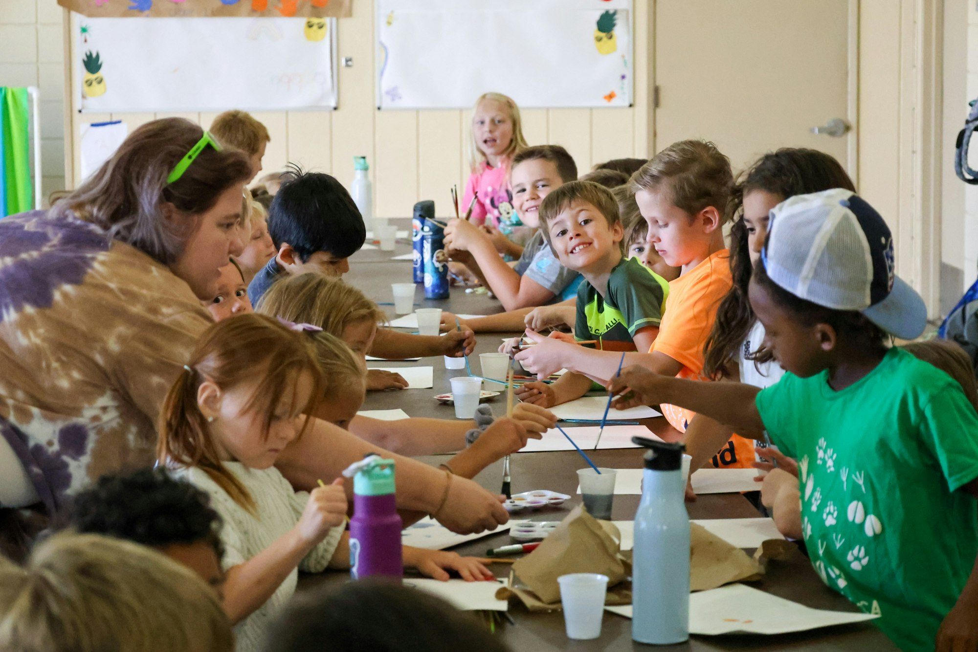 Children engaged in a group art activity, painting and drawing at a long table.