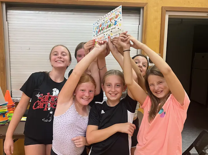 A group of happy girls hold up a colorful thank-you sign, celebrating together in a cozy indoor setting.