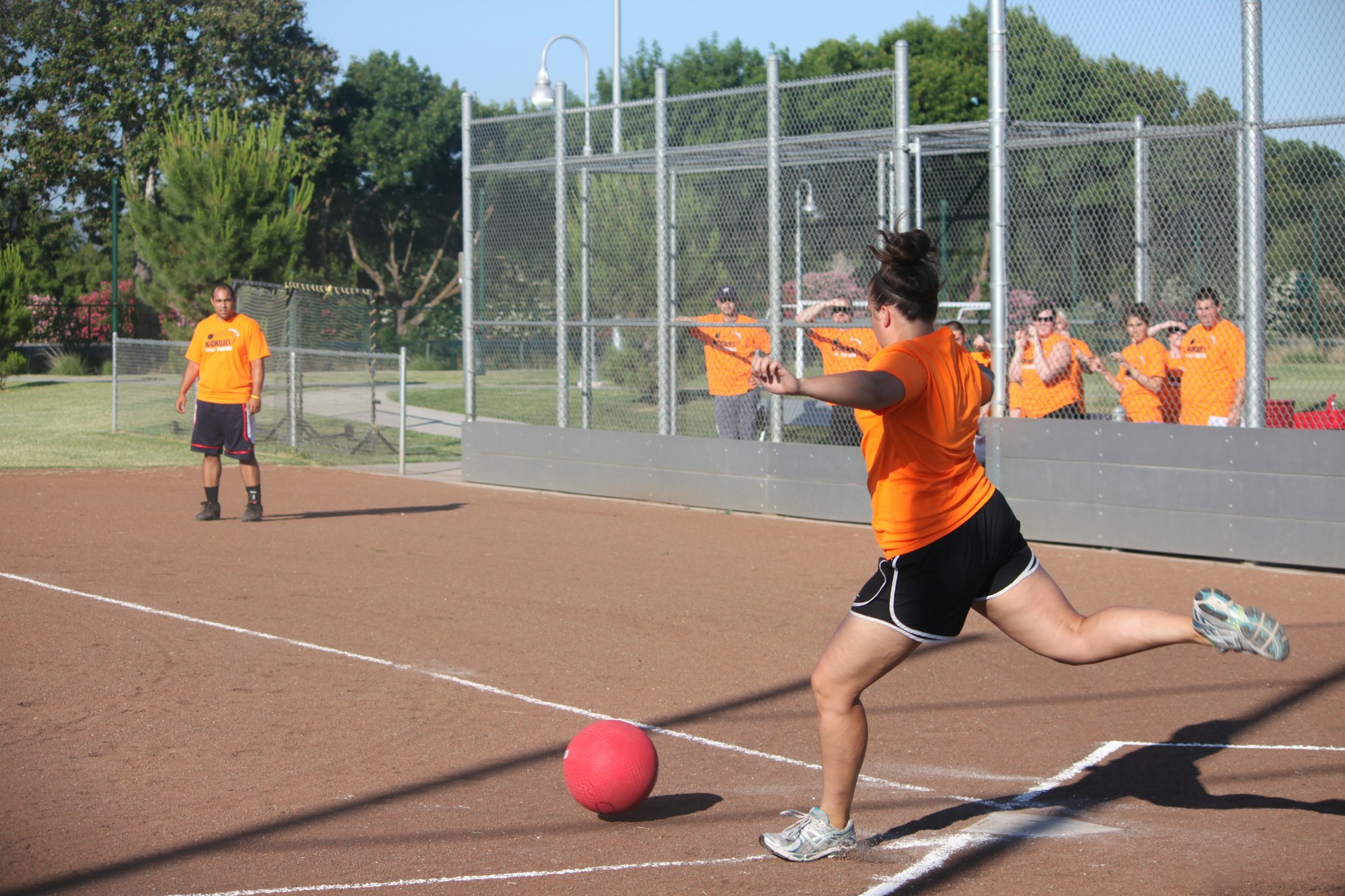 A kickball game in progress with players in orange shirts. One athlete is kicking the ball while others watch from the sidelines.