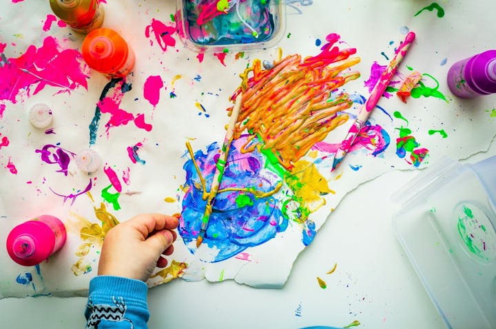 Colorful abstract painting with brushes, paint bottles, and a child's hand on messy paper.