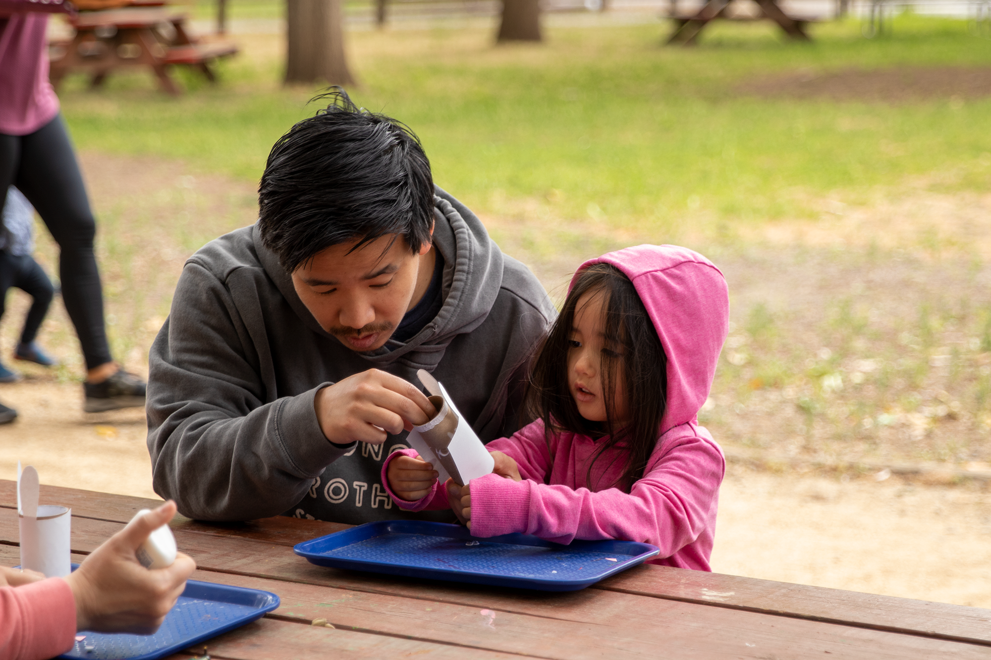 A man and a young girl work together at a table, crafting with materials, enjoying a moment outdoors.