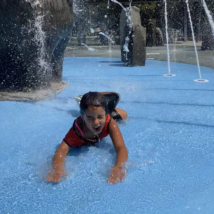 A child joyfully slides on a blue surface, splashing water around from fun fountains in a sunny setting.
