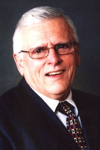 An elderly man wearing glasses, a dark suit, and a patterned tie, smiling at the camera against a dark background.
