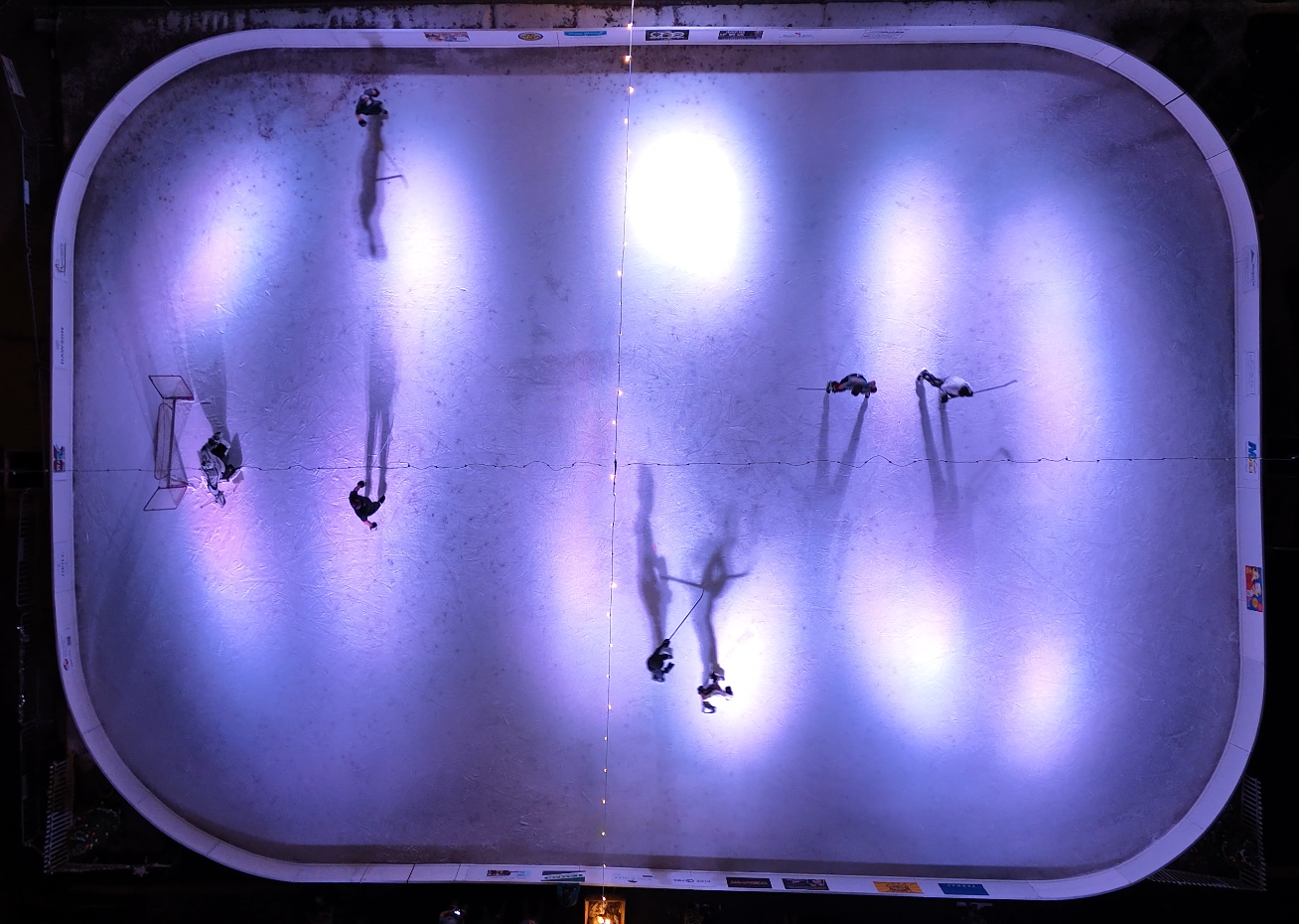 Overhead view of an illuminated ice rink with people playing hockey, casting long shadows on the ice.